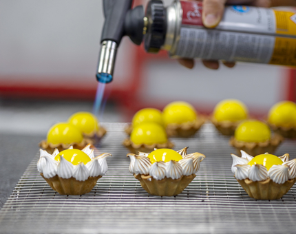 A hand holding a catering blow torch is firing meringues on small yellow cakes. 