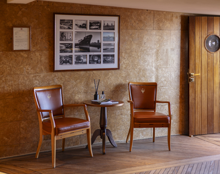 The reception area of Fingal Hotel. There are two wooden chairs covered in brown leather, a side table between the, and a photo frame on the wall with black and white photos of the ship.