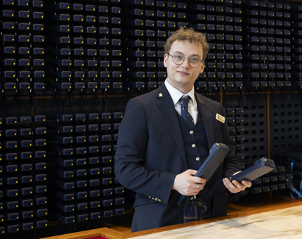 A man is posing for a photo at Britannia's handset desk, behind him is a wall where all of the handsets are stored. 