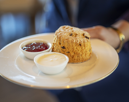 A hand holding a white plate with a fruit scone and jam and cream. 