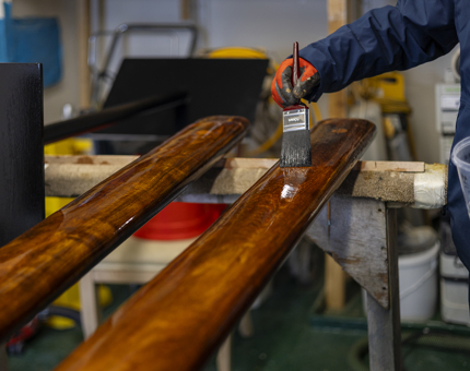 A close up of a hand holding a paint brush applying varnish to wooden handrails. 