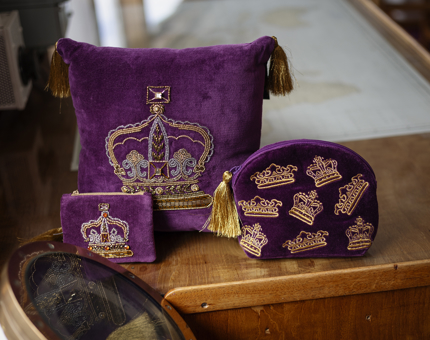 The crown range of merchandise from the Britannia Shop. There is a purple cushion, and two makeup bags with a gold crown design on them. 