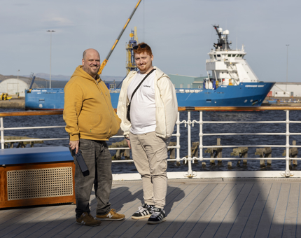 Two men posing for a photo on the Verandah Deck. There is a large blue and white ship in the background. 
