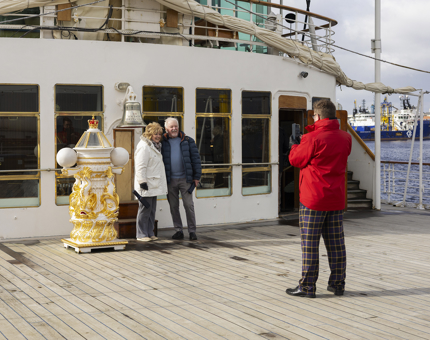 A man wearing a red jacket is taking a photo of two people at Britannia's Bell on the Verandah Deck. 