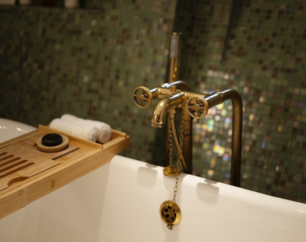 A close up of a bath with brass fittings. The wall behind it has green tiles. 