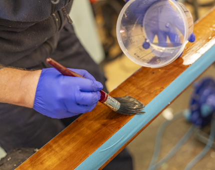 A close up of a hand holding a plastic tub and paintbrush, varnishing a handrail. 