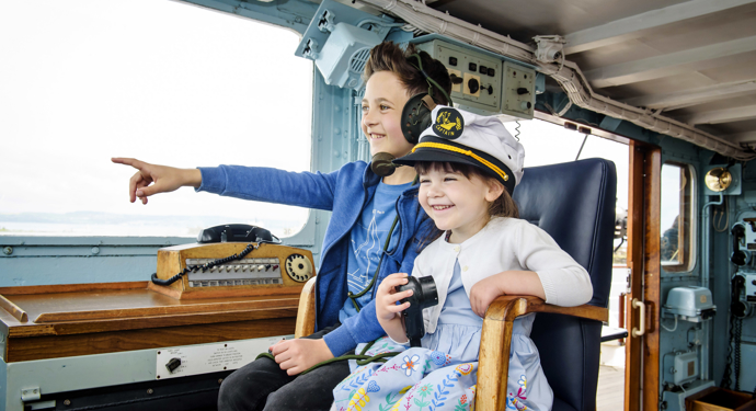 Two children sitting on the Captain's chair in the Bridge. The girl is wearing a Captain's hat, the boy is wearing a headset. 