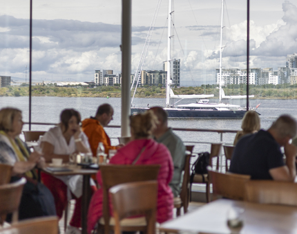 Visitors sitting at tables in the Royal Deck Tearoom. The floor to ceiling windows in the background have a view of a yacht sailing into the port.