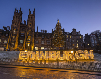 The word Edinburgh made up of lights. There are historic buildings in the background. 