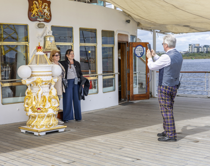 A man is taking a photo of visitors next to the gold and white binnacle on Britannia's Verandah Deck in the Port of Leith. 