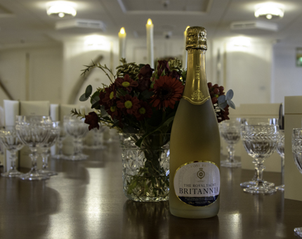 A bottle of Britannia's Sparkling Rosé sits next to a vase of red flowers on a table within the State Dining Room.