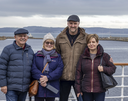 A family group of four people stand against the Bridge with a seaview background.