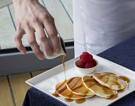 A Chef pouring a small jug of syrup over a plate of pancakes, There is a small dish of raspberries on the plate too.
