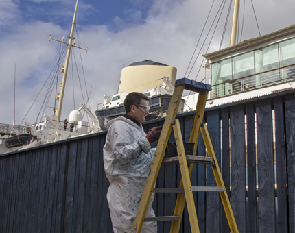 A Facilities team member standing on a ladder painting a fence with blue paint.