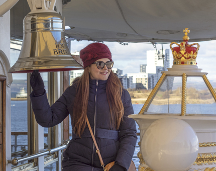A woman visitor wearing a jacket and hat standing next to the Bell on the Verandah Deck. She is pretending to ring the bell.