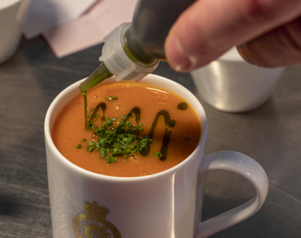 A finishing drizzle is added to a mug of tomato and red pepper soup.