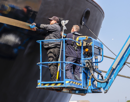 Two members of the Maintenance team on a cherrypicker painting the side of Britannia.