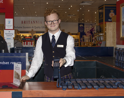 A smiling man holding an audio guide handset standing at a desk where the audio guides are given to visitors.