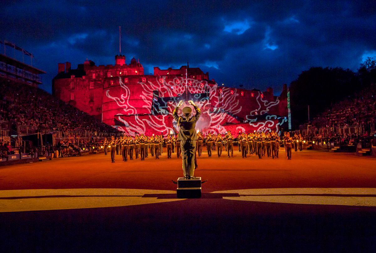 Edinburgh Castle is lit up with red lights, there is a man conducting a marching band at the Royal Edinburgh Military Tattoo.