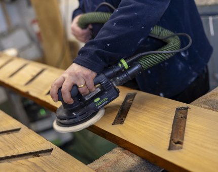 A close up of a hand holding an electric sander to sand a wooden ladder. 
