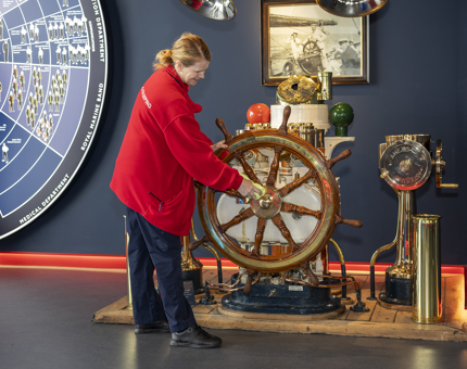 A Housekeeper wearing a red fleece is in the Visitor Centre in Leith cleaning the ship's wheel. 