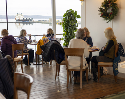 People are sitting at tables in the Royal Deck Tearoom, through the windows in the background you can see a ship in the Port of Leith. 