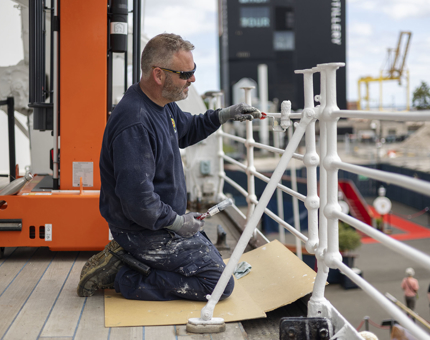 A Maintenance team member holding a paint brush and kneeling next to the railings on deck, painting them white.