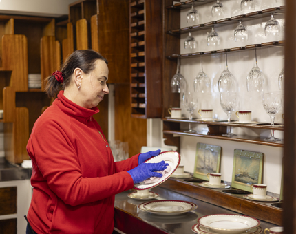 A Housekeeper wearing a red fleece is polishing a plate in the china pantry with a cloth.