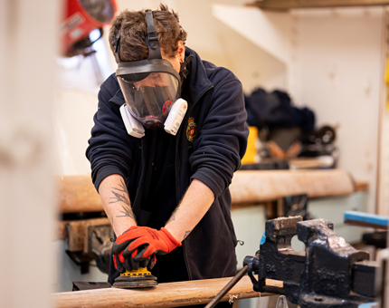 In the Joiner's workshop, a Maintenance man is using both hands to hold a sander to sand varnish off of a wooden handrail. He is wearing a full face protection mask. 