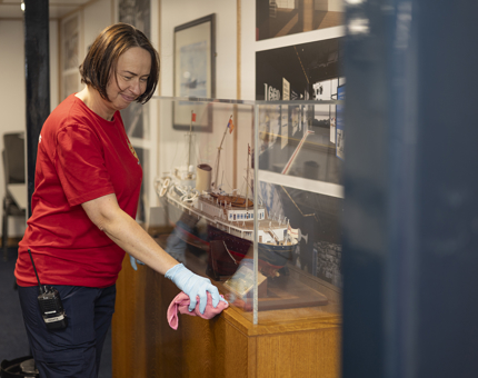 A Housekeeper is holding a pink cloth, wiping down a glass case containing a model of The Royal Yacht Britannia. 