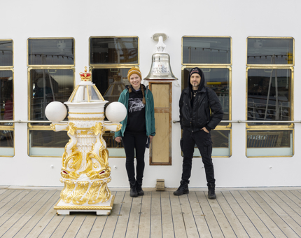 A man and woman standing on the Verandah Deck of Britannia in Leith by the ship's Bell and gold and white decorative binnacle. 