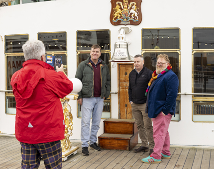 A man wearing a red jacket is taking a photo of three men posing by the Bell on The Royal Yacht Britannia's Verandah Deck. 