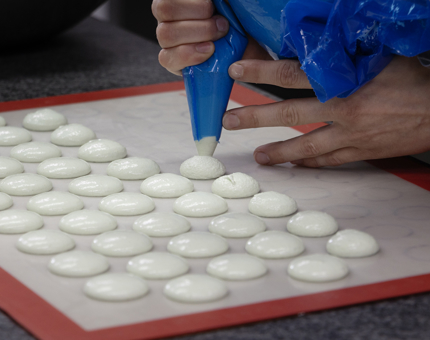 A close up of a Chef using a piping bag to pipe macaron batter onto a tray. 