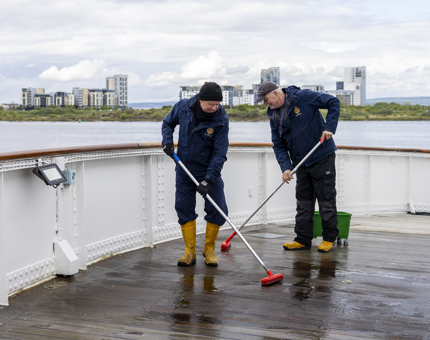 Two Maintenance team members using brooms to scrub the deck of Britannia in the Port of Leith. 