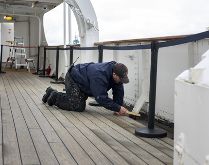 A Maintenance man kneeling on the deck, he is using a wire brush to remove paint. 