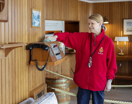 A Housekeeper is wearing a red fleece and is wiping a wooden telephone box attached to the wood paneled wall of the Sun Lounge aboard The Royal Yacht Britannia.   