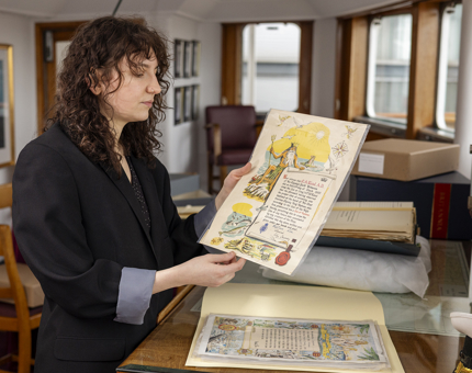 A woman is showing a colourful illustrated document to the camera in the Chartroom aboard Britannia in Leith. 