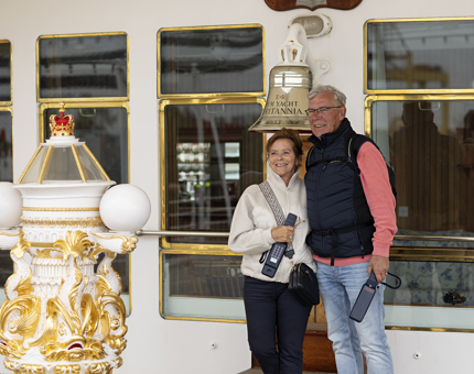 A man and a woman holding audio guide handsets pose for a photo next to Britannia's Bell and decorative binnacle. 