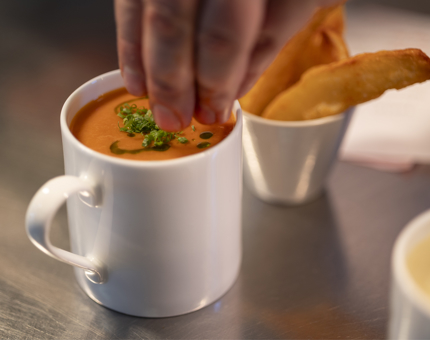 A hand is sprinkling shives onto a mug of red pepper soup. there is a bowl of potato wedges behind it. 