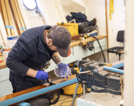 A maintenance team member holding a tube and a paint brush, applying a coat of varnish to a handrail. 