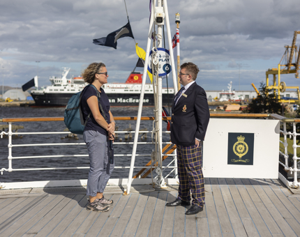 On The Royal Yacht Britannia in the Port of Leith, two people are talking on the deck, there is a ship in the background. 