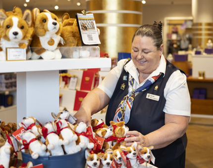 A Sales Assistant is in Britannia's Shop, restocking the plush corgi toys. 