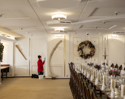 A Housekeeper polishing brass in Britannia's State Dining Room where a long table is set and a Christmas wreath hangs on the far wall. 