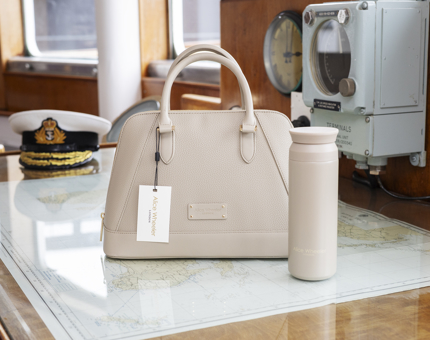 An Alice Wheeler Stone Metal Water Bottle And Kensington Handbag sit on a bench which has a map on it, there is a captain's hat in the background. 