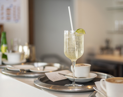 A tray with a cocktail with a white straw and lime wedge. There are also tray the have coffees on them.