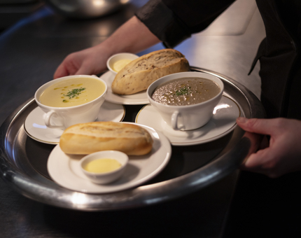 Hands are holding a round tray with two bowls of soup and two plates with rolls and butter on them in the Royal Deck Tearoom. 