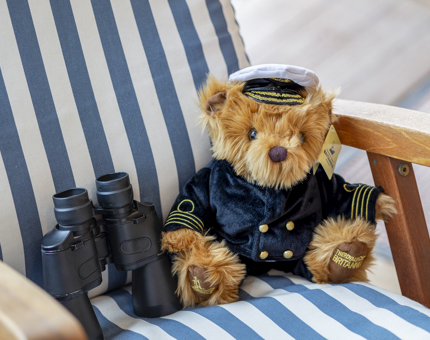 A captain plush bear is sitting on a blue and white striped seat with a pair of binoculars next to it.