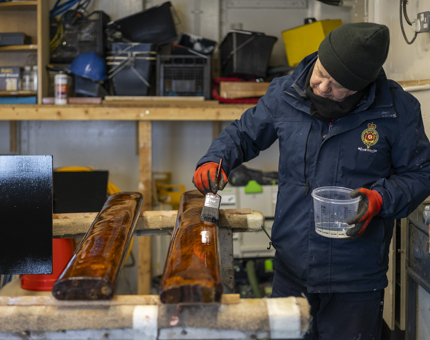 A Maintenance man is holding a tub of varnish and a paint brush, applying varnish to wooden handrails. 