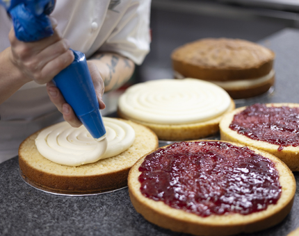 A close-up of a chef piping cream onto cake bases. There are other cake bases with jam on them. 