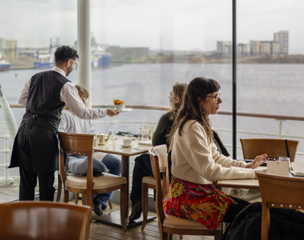 A server is placing a bowl of potato wedges on a table in the Royal Deck Tearoom aboard Britannia. There is a view of the Port of Leith through the window. 